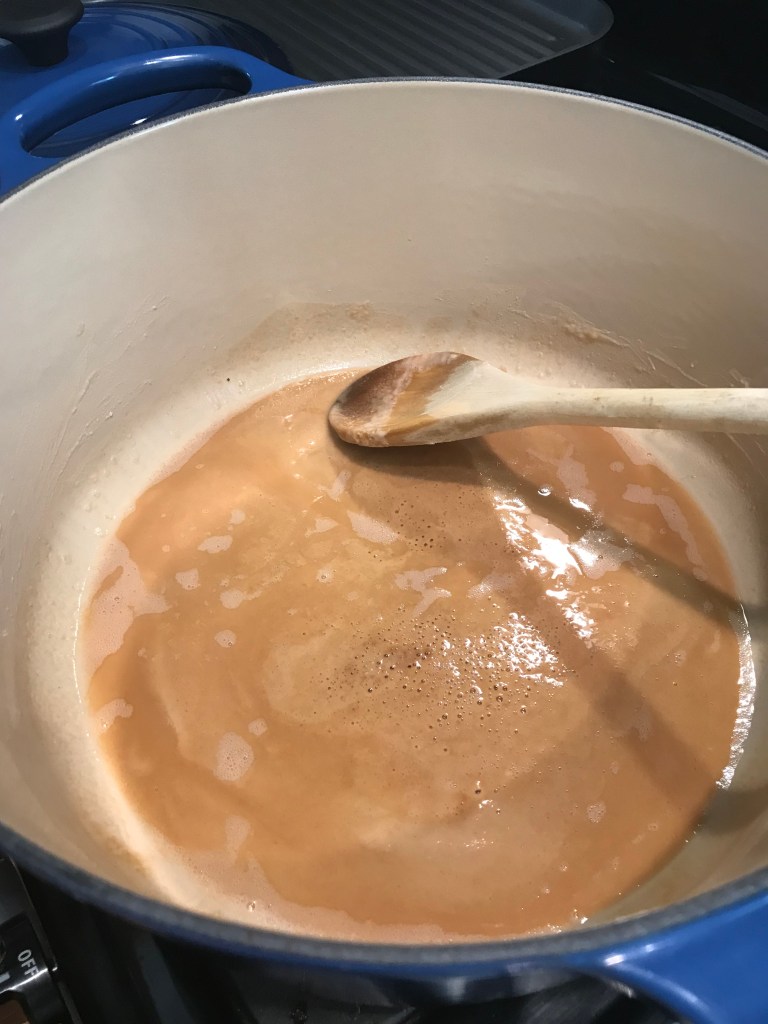 A pot containing a light-colored roux with a wooden spoon stirring it, showing a smooth, bubbling mixture.