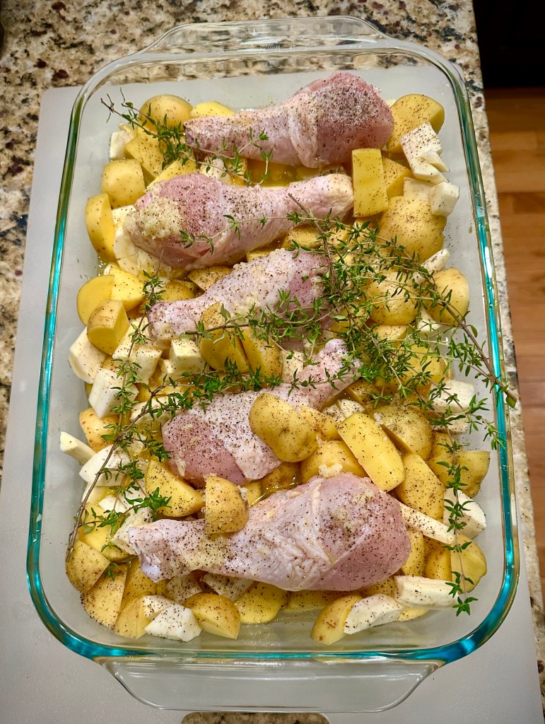 A glass baking dish filled with seasoned chicken drumsticks on a bed of halved baby Yukon Gold potatoes and chopped parsnips, garnished with fresh thyme.