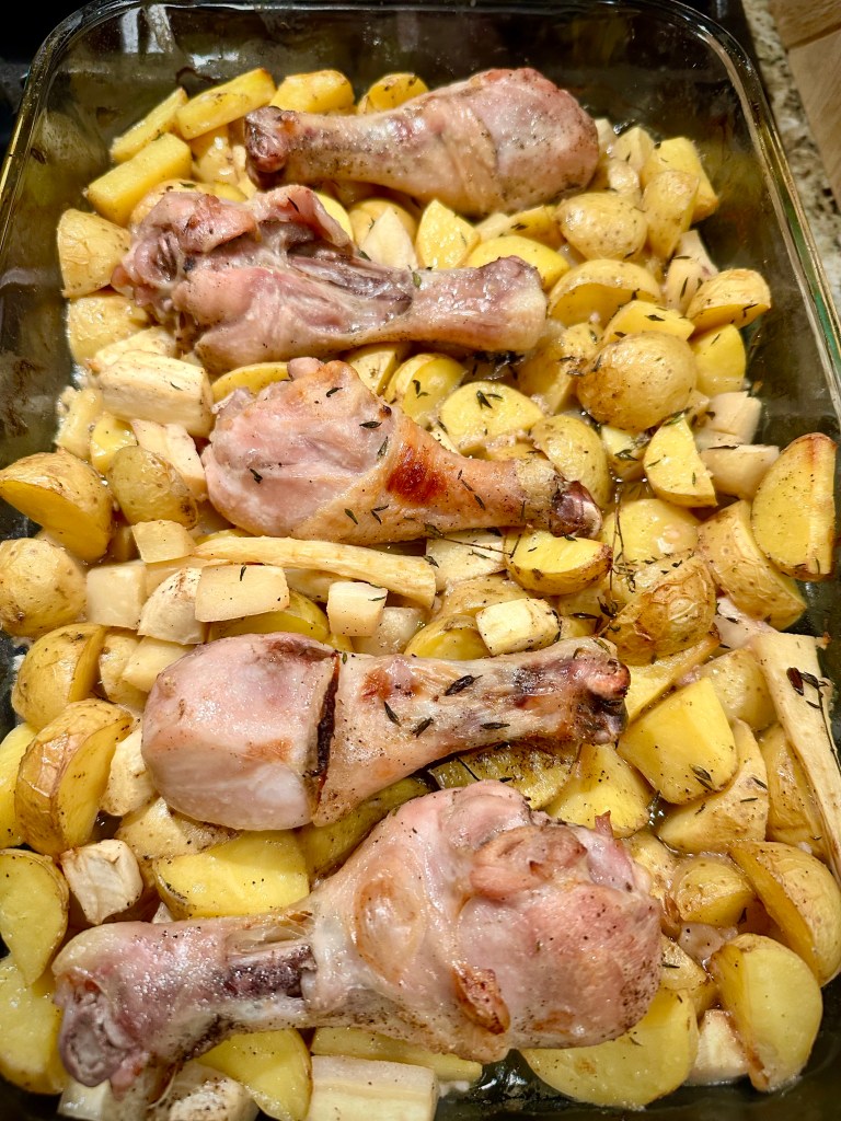 A glass baking dish filled with cooked chicken drumsticks placed on a bed of halved baby Yukon Gold potatoes and chopped parsnips, seasoned with herbs.