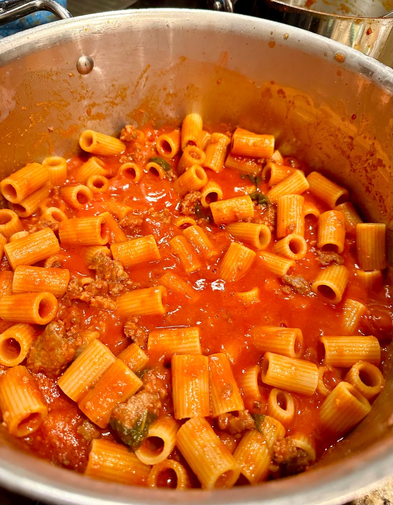 A pot filled with rigatoni pasta mixed with Italian sausage in a rich tomato sauce, garnished with fresh herbs.