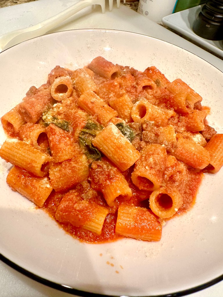 A bowl of rigatoni pasta topped with a rich tomato sauce, sprinkled with grated Pecorino Romano cheese and garnished with fresh basil.