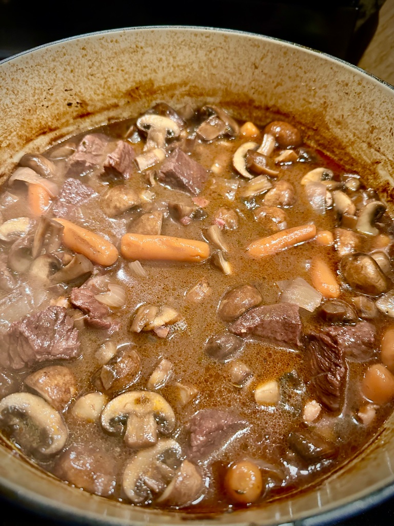 A pot of beef stew with chunks of beef, mushrooms, and baby carrots simmering in rich brown sauce.