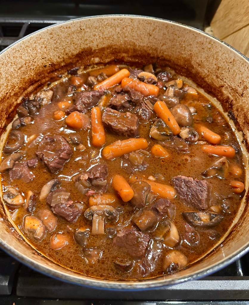 A pot of beef stew featuring chunks of tender beef, orange baby carrots, and whole mushrooms cooked in a rich red wine sauce.